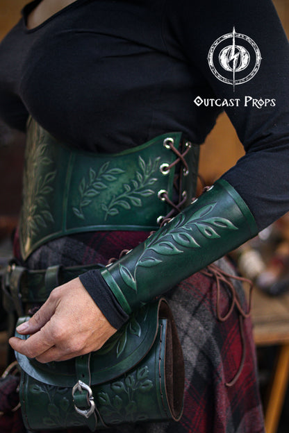 Side view of a woman wearing a green leaf elven bracer with a matching corset and hip bag decorated with embossed leaf patterns. Ideal for renaissance faire fashion, larp characters, elves, witches, herbalists, nobles and fae performers. The leaf motif also suits roman laurel or Caesar themed cosplay. Soft lighting and rustic textures highlight the hand aged leather and elegant fantasy craftsmanship of the ensemble.