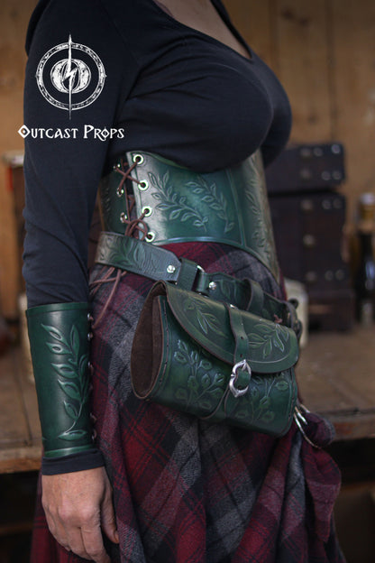 A full outfit view shows a woman wearing a green leaf elven bracer, matching corset and embossed leather hip bag. The coordinated leaf motifs create a refined renaissance faire look for elves, witches, herbalists, nobles and fae characters. This leather larp bracer also suits roman laurel or Caesar cosplay thanks to its classic leaf design. The rustic background enhances the fantasy themed craftsmanship and aged textures.