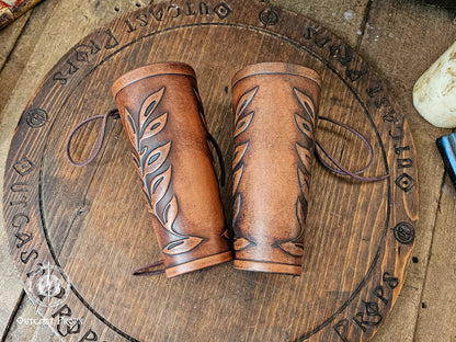 Two matching brown leaf elven bracers displayed on a carved wooden board with the Outcast Props logo. Their embossed leaf pattern and aged leather finish show craftsmanship ideal for renaissance faire costumes, larp characters, elves, witches, herbalists or fae themed outfits. These leather bracers also complement roman laurel or Caesar cosplay thanks to the leaf motif. Laced fastening and warm tones highlight their elegant look.
