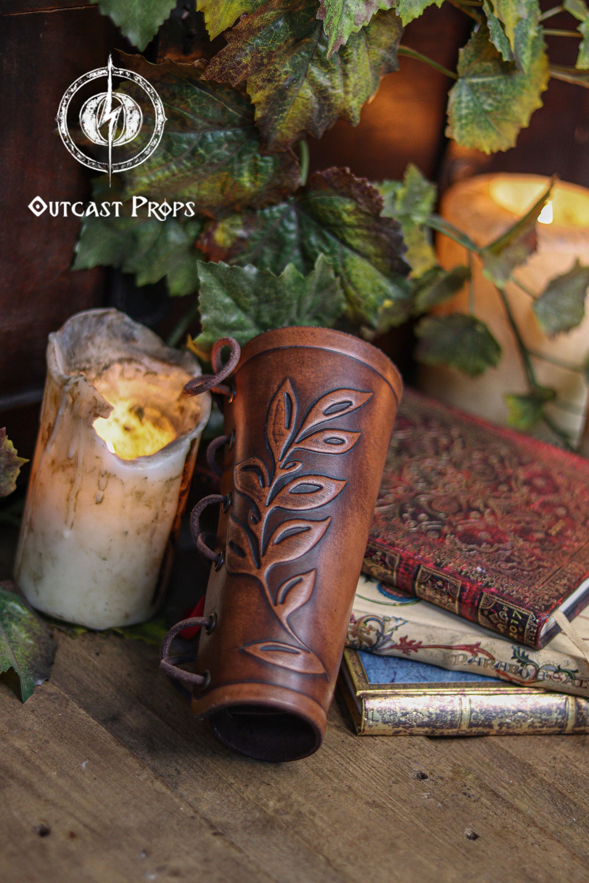 Close up of a brown leaf elven bracer with embossed leaf details, displayed beside vintage books, a worn candle and green foliage. The natural curve and laced fastening show its elegant design for renaissance faire outfits, larp costumes and cosplay characters like elves, witches, herbalists or fae performers. Made of hand dyed leather, it also suits roman laurel or Caesar themed looks. Rustic lighting enhances the handcrafted textures.