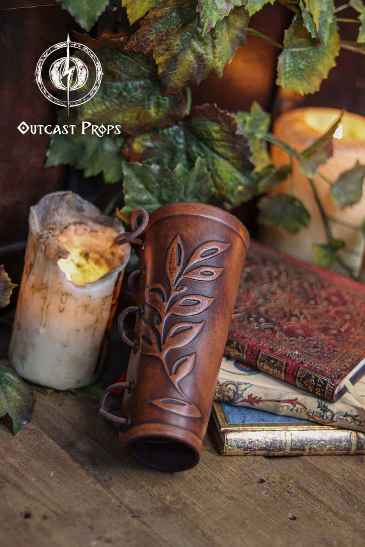 Close up of a brown leaf elven bracer with embossed leaf details, displayed beside vintage books, a worn candle and green foliage. The natural curve and laced fastening show its elegant design for renaissance faire outfits, larp costumes and cosplay characters like elves, witches, herbalists or fae performers. Made of hand dyed leather, it also suits roman laurel or Caesar themed looks. Rustic lighting enhances the handcrafted textures.