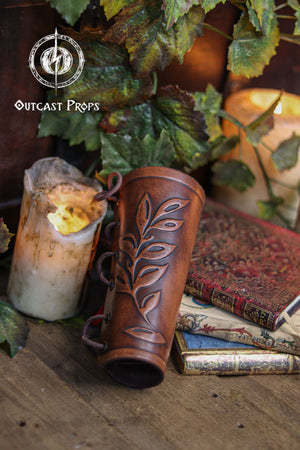 Close up of a brown leaf elven bracer with embossed leaf details, displayed beside vintage books, a worn candle and green foliage. The natural curve and laced fastening show its elegant design for renaissance faire outfits, larp costumes and cosplay characters like elves, witches, herbalists or fae performers. Made of hand dyed leather, it also suits roman laurel or Caesar themed looks. Rustic lighting enhances the handcrafted textures.