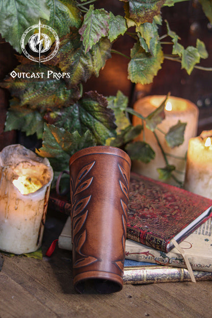 A brown leather leaf elven bracer lies on top of antique books near warm candles and green ivy, creating a renaissance faire atmosphere. The embossed leaf pattern adds elegance for elves, witches, herbalists, nobles or fae costumes. This larp leather bracer works beautifully for roman laurel or Caesar inspired cosplay thanks to its classical leaf design. Hand aged surface and laced closure complete this fantasy accessory.