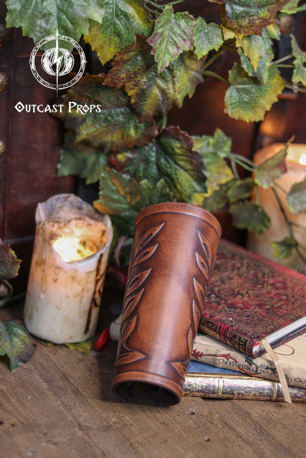A brown leather leaf elven bracer lies on top of antique books near warm candles and green ivy, creating a renaissance faire atmosphere. The embossed leaf pattern adds elegance for elves, witches, herbalists, nobles or fae costumes. This larp leather bracer works beautifully for roman laurel or Caesar inspired cosplay thanks to its classical leaf design. Hand aged surface and laced closure complete this fantasy accessory.