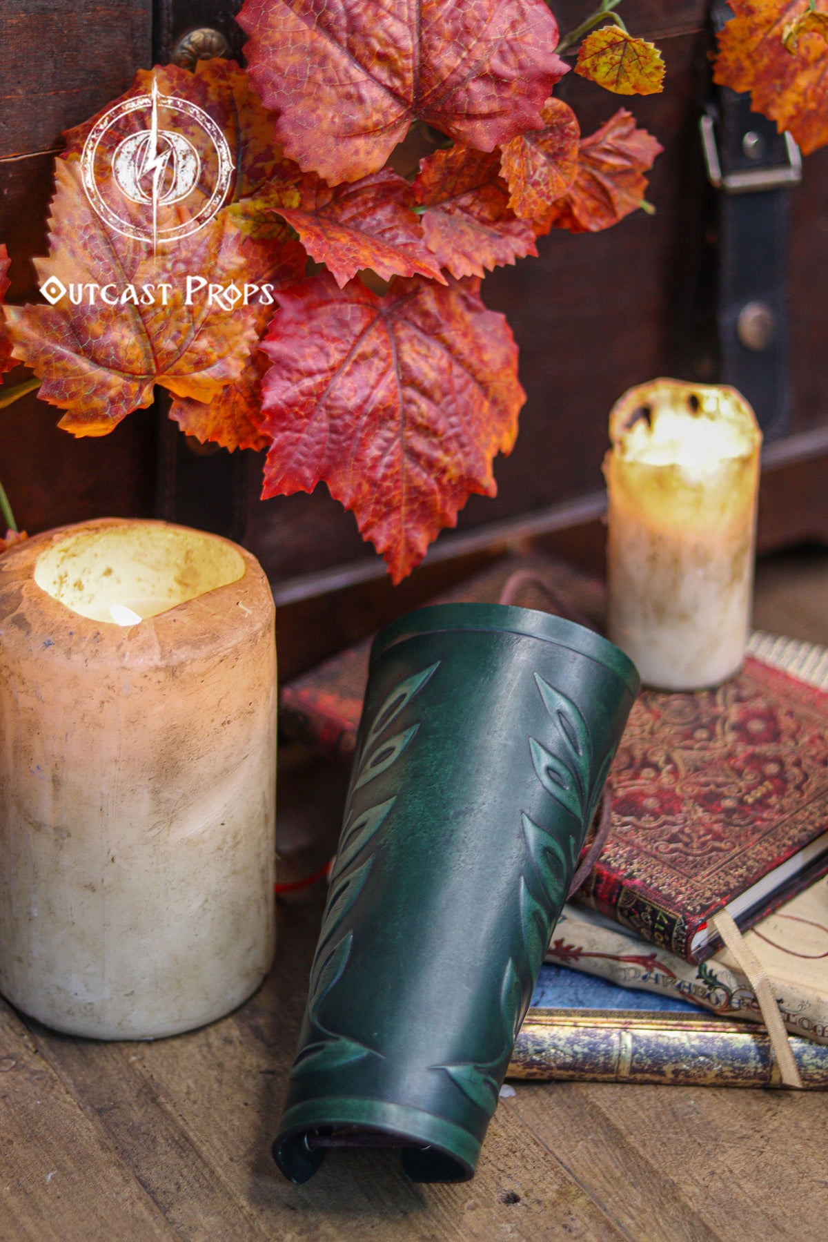 A green leaf elven bracer rests beside glowing candles, red autumn leaves and antique books. The embossed leaf motif and deep forest color make it perfect for renaissance faire costumes, elves, witches, herbalists, fae characters and larp outfits. Its classic leaf design also suits roman laurel or Caesar cosplay. The warm rustic scene highlights the hand aged leather and elegant fantasy style of this versatile accessory.