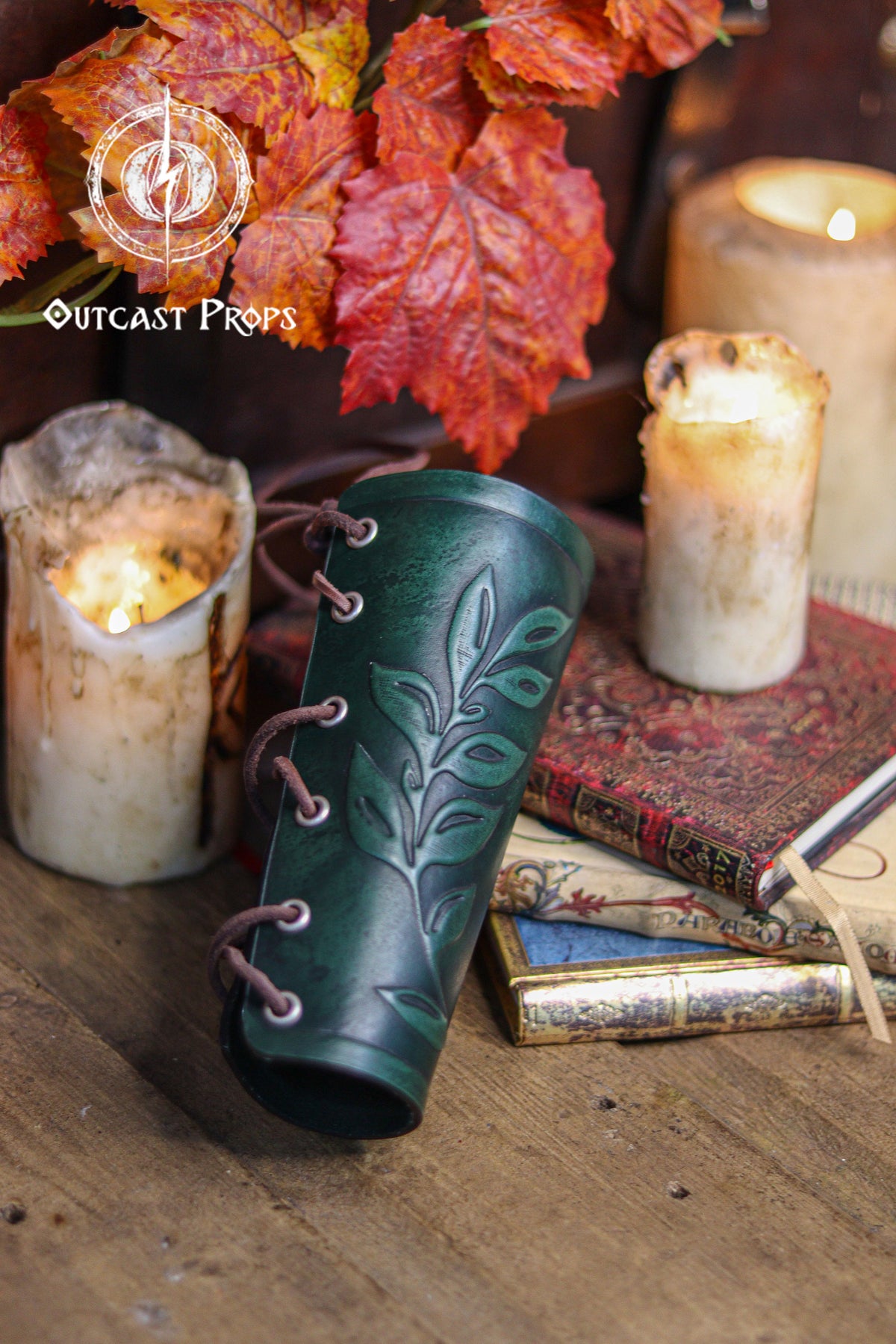 A green leaf elven bracer stands upright beside warm candles, autumn foliage and vintage books in a renaissance faire styled setting. The embossed leaf pattern and laced closure highlight its handcrafted design for elves, witches, herbalists, nobles and fae performers. This leather larp bracer also works beautifully for roman laurel or Caesar themed cosplay. Textured lighting enhances its fantasy aesthetic and elegant curved silhouette.