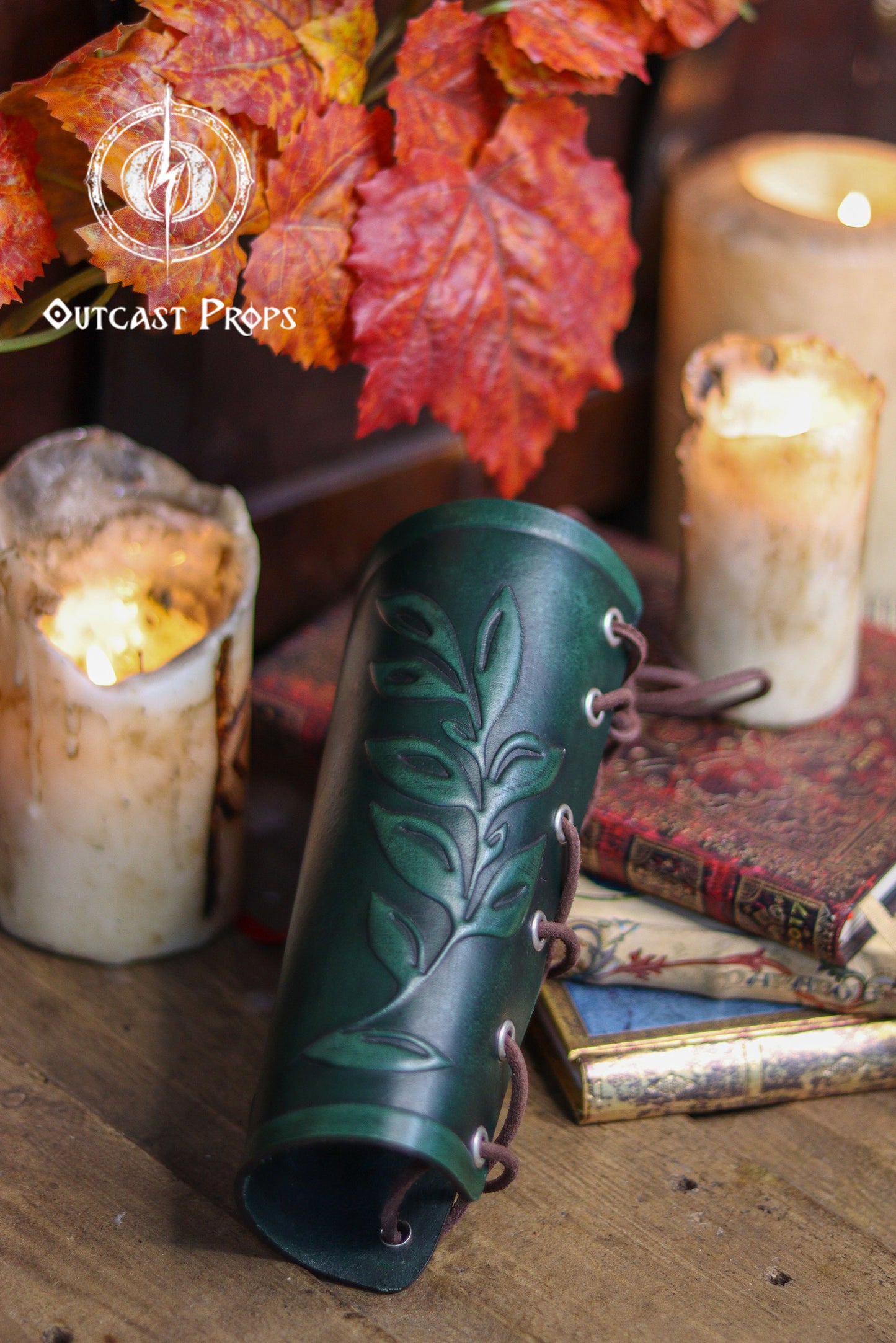 A green leaf elven bracer lies across antique books near softly lit candles and orange autumn leaves. Its embossed leaf design and aged leather finish make it ideal for renaissance faire outfits, elves, witches, herbalists, fae characters and larp costumes. The classical leaf motif also suits roman laurel or Caesar cosplay. Warm tones and rustic textures highlight the craftsmanship and natural fantasy look of the accessory.