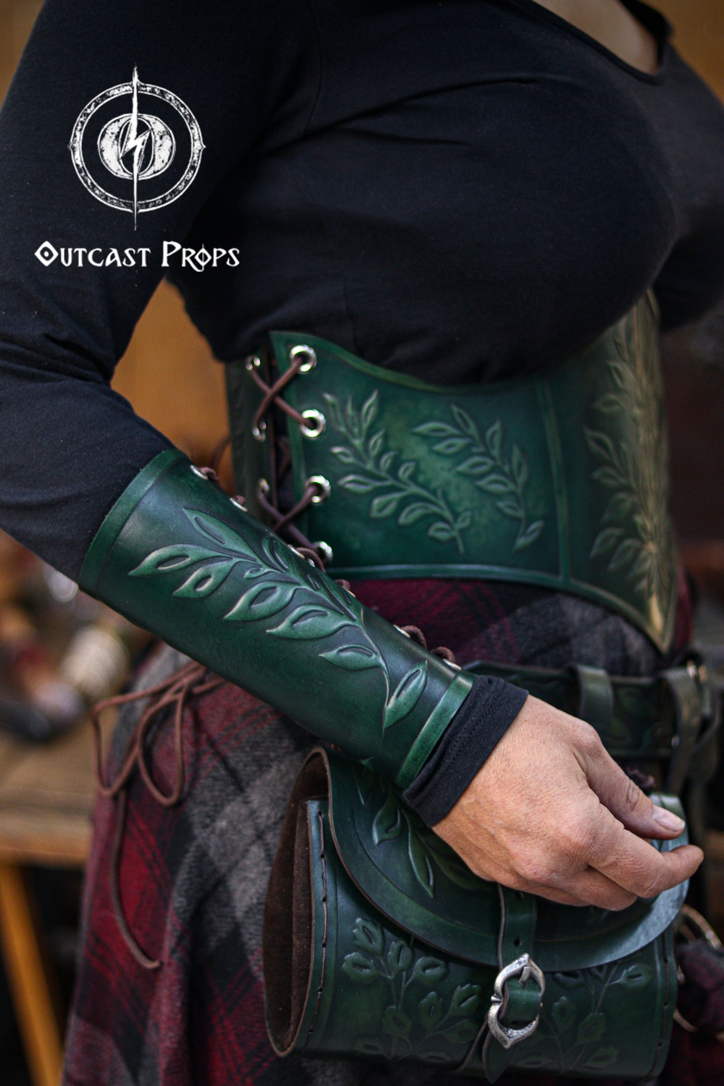 A woman wearing a green leaf elven bracer poses with a matching embossed leather corset and hip bag. The leaf motif creates a cohesive renaissance faire outfit perfect for elves, witches, herbalists, nobles, fae performers and larp characters. The bracer’s shape also complements roman laurel or Caesar inspired cosplay. The scene highlights hand aged leather, laced details and the elegant fantasy style of the set.