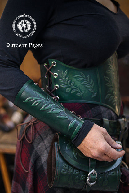 A woman wearing a green leaf elven bracer poses with a matching embossed leather corset and hip bag. The leaf motif creates a cohesive renaissance faire outfit perfect for elves, witches, herbalists, nobles, fae performers and larp characters. The bracer’s shape also complements roman laurel or Caesar inspired cosplay. The scene highlights hand aged leather, laced details and the elegant fantasy style of the set.