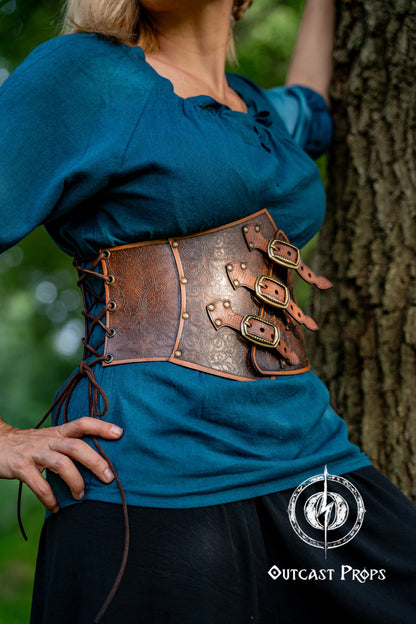 Person wearing a blue top and brown leather corset with decorative elements, standing against a tree. Embroidered Leather Underbust Corset – Elegant Brown Waist Cincher for Noble LARP Outfits, Steampunk and Fantasy Glamour