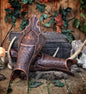 Artisanal Celtic greaves displayed on a wooden stage surrounded by Viking props, antlers, and rune candles. The brown leather leg armor features Nordic knotwork and a dragon design. Handmade by Outcast Props for fantasy warriors, LARP adventurers, or medieval cosplay outfits.