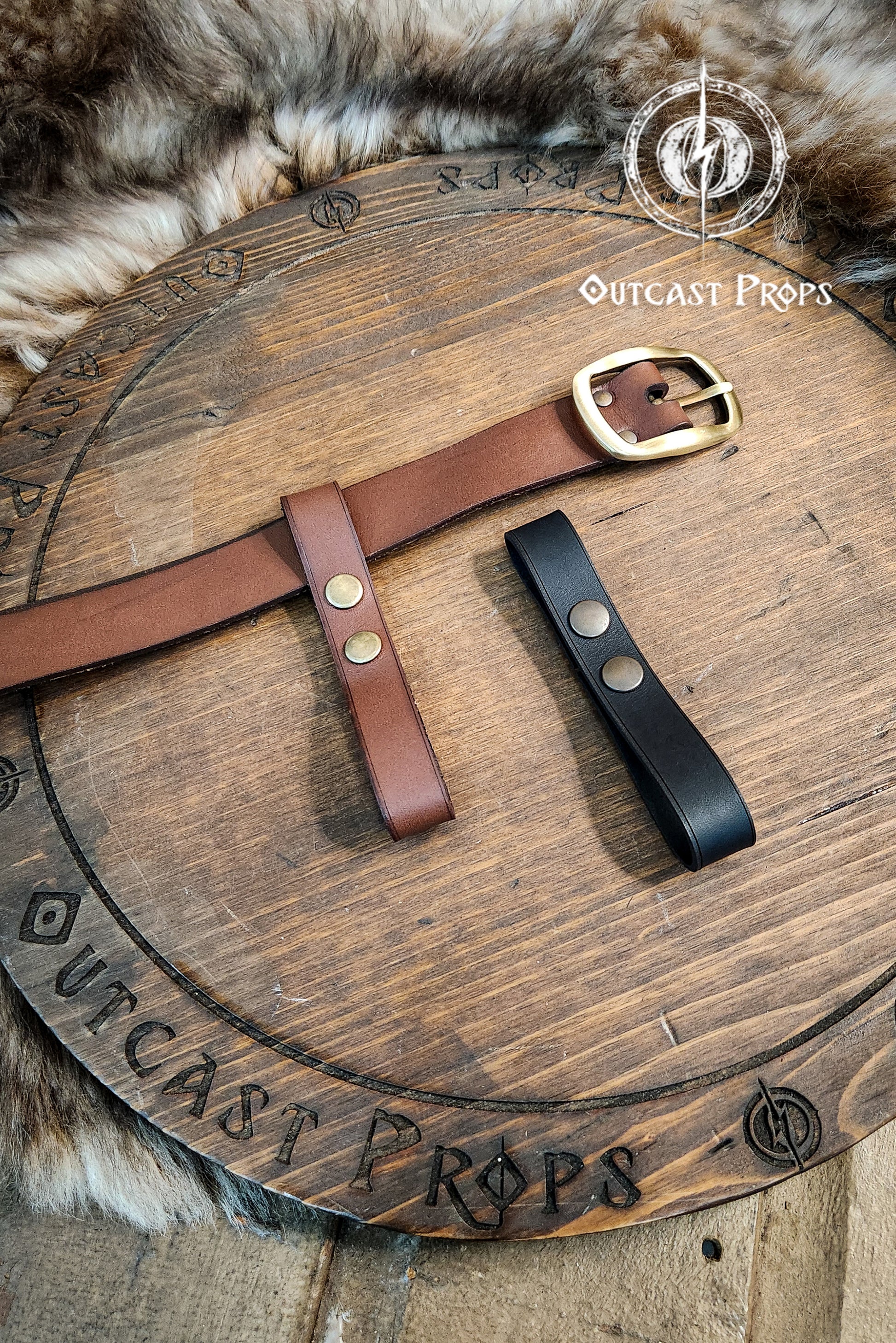 Two leather belts, one brown and one black, on a wooden surface with 'Outcast Props' branding. Versatile Leather Belt Clasp for LARP, Cosplay & Medieval Accessories - Handcrafted Vegetal Tanned Attachment