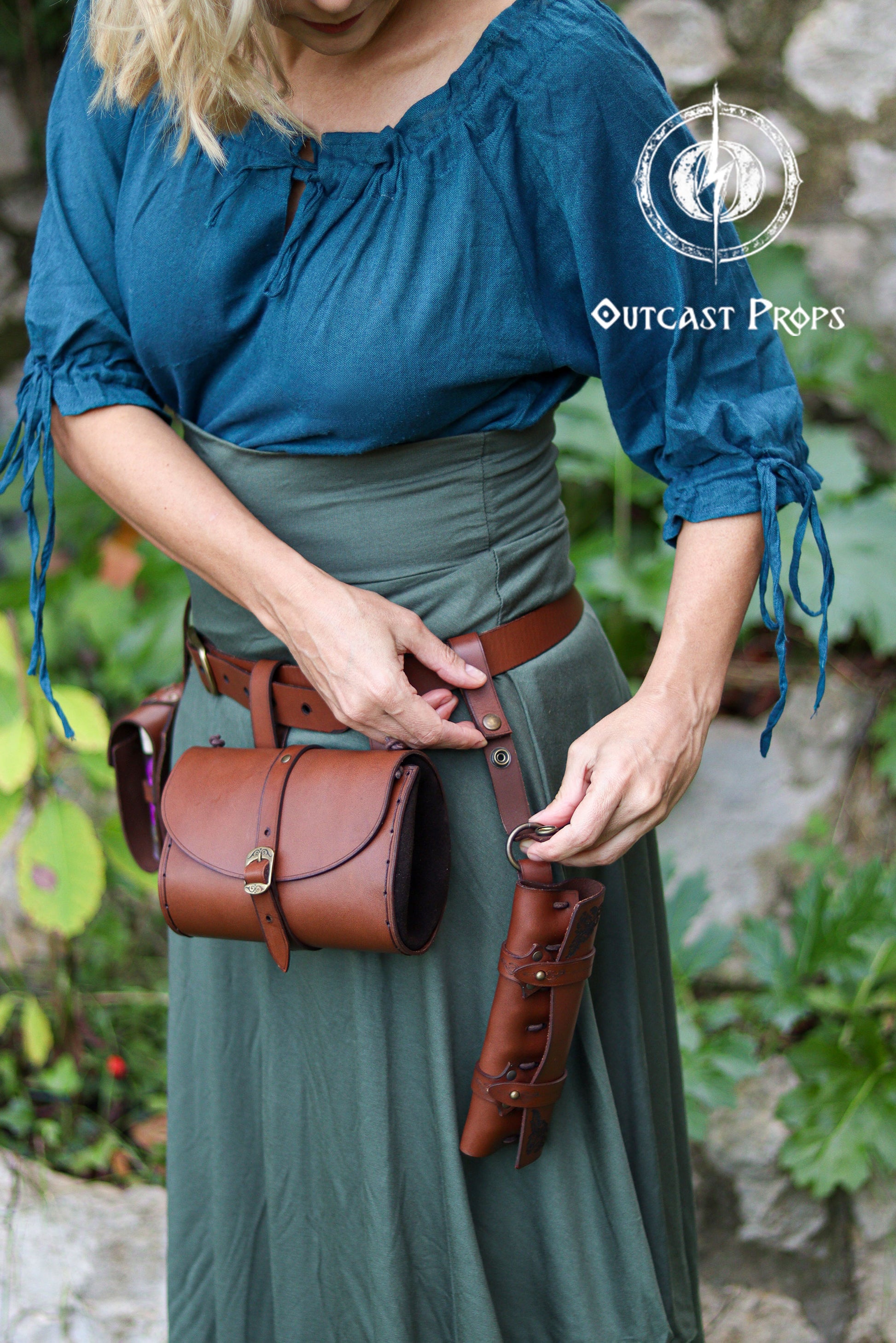 A blonde woman adjusts a leather clasp belt strap attached to her waist belt, styled in teal and forest green clothing reminiscent of fantasy herbalists or adventurers. The clasp features strong snaps and a metal ring for hanging items such as pouches, mug holders, scabbards or potion holsters. Shown paired with a cylindrical pouch and a multi-vial potion case, the accessory enhances LARP, steampunk and Renaissance Faire costumes by adding modular carrying options without extra bulk.