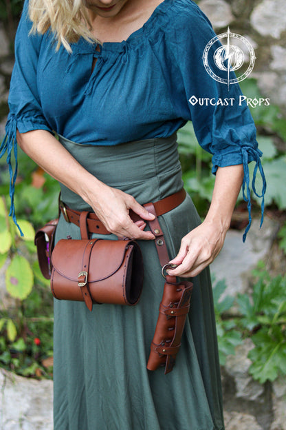 A blonde woman adjusts a leather clasp belt strap attached to her waist belt, styled in teal and forest green clothing reminiscent of fantasy herbalists or adventurers. The clasp features strong snaps and a metal ring for hanging items such as pouches, mug holders, scabbards or potion holsters. Shown paired with a cylindrical pouch and a multi-vial potion case, the accessory enhances LARP, steampunk and Renaissance Faire costumes by adding modular carrying options without extra bulk.