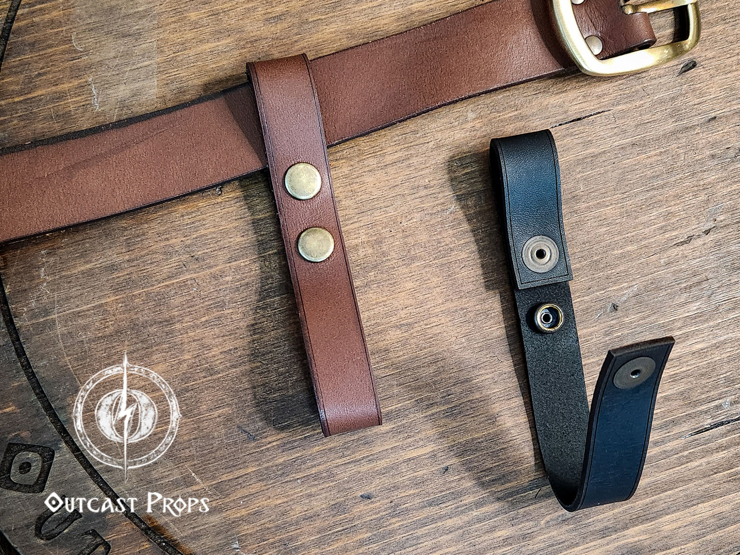 Brown and black leather belts with brass buckles on a wooden surface, featuring Outcast Props logo. Versatile Leather Belt Clasp for LARP, Cosplay & Medieval Accessories - Handcrafted Vegetal Tanned Attachment