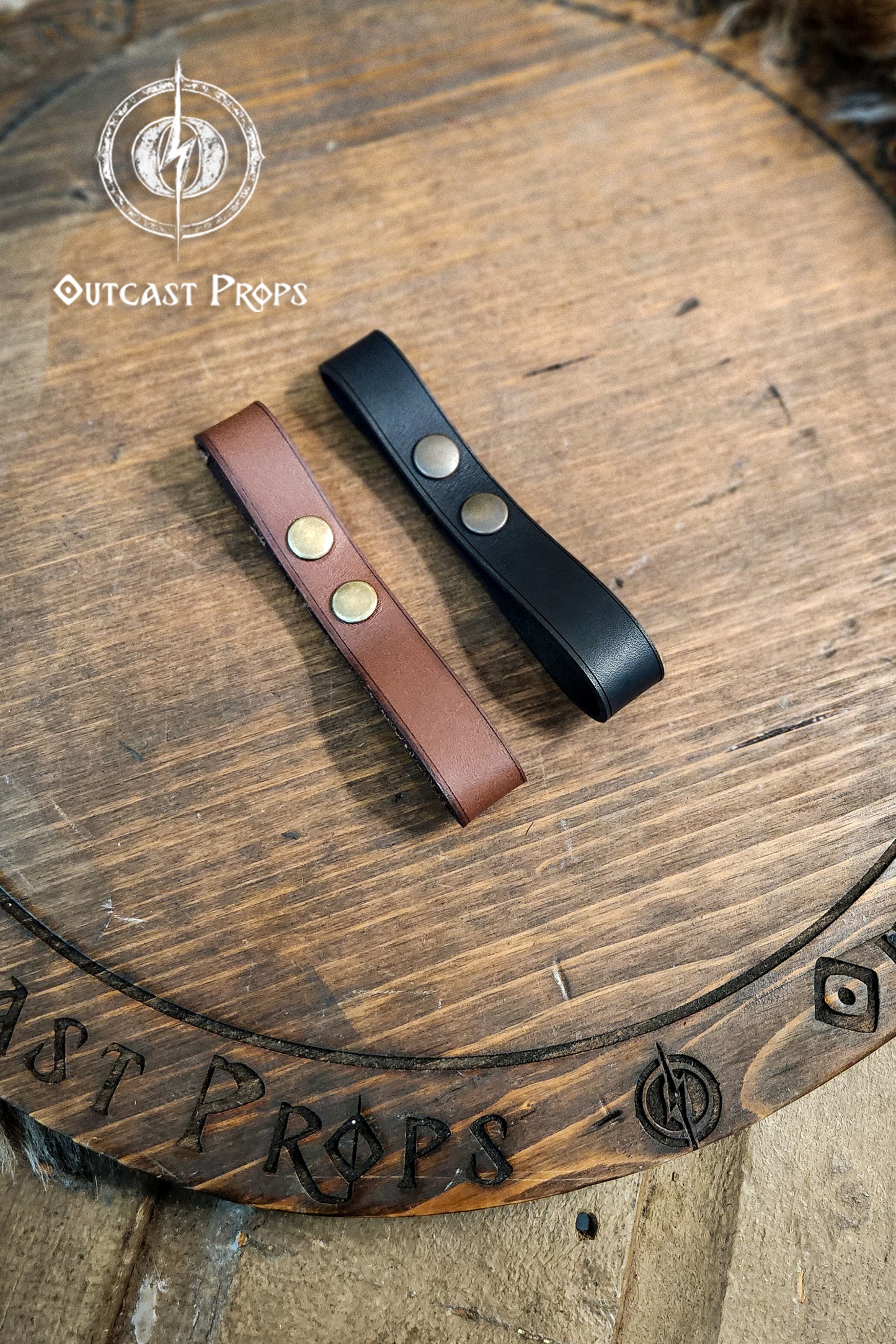 Two leather straps, one brown and one black, on a wooden surface with 'Outcast Props' branding. Versatile Leather Belt Clasp for LARP, Cosplay & Medieval Accessories - Handcrafted Vegetal Tanned Attachment