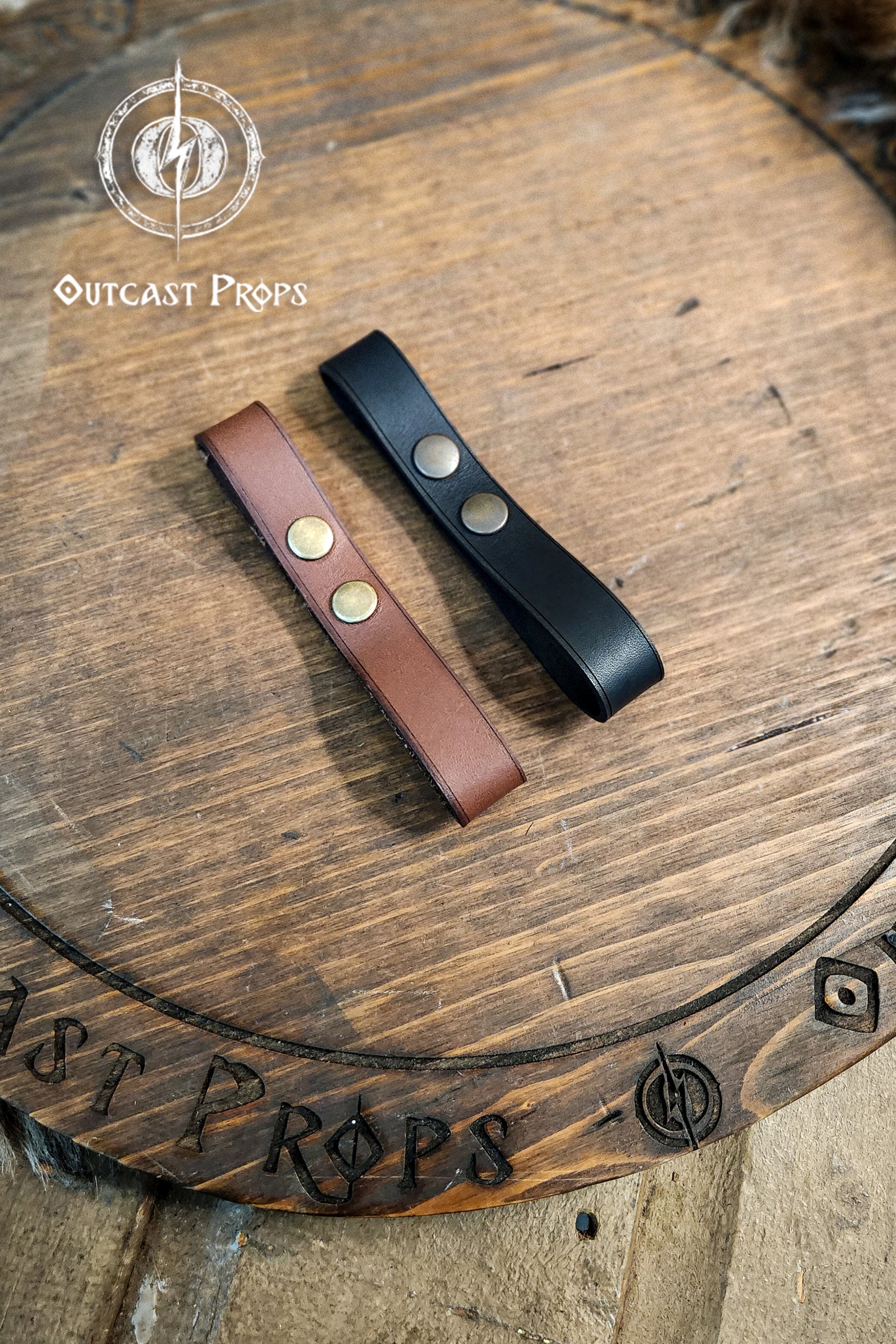 Two leather straps, one brown and one black, on a wooden surface with 'Outcast Props' branding. Versatile Leather Belt Clasp for LARP, Cosplay & Medieval Accessories - Handcrafted Vegetal Tanned Attachment
