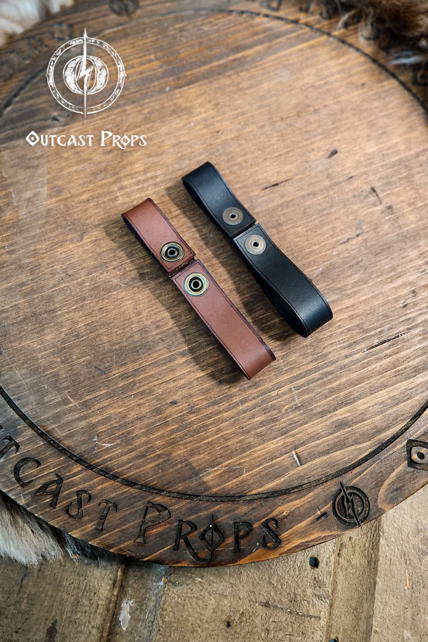 Two leather straps, one brown and one black, on a wooden surface with 'Outcast Props' branding. Versatile Leather Belt Clasp for LARP, Cosplay & Medieval Accessories - Handcrafted Vegetal Tanned Attachment