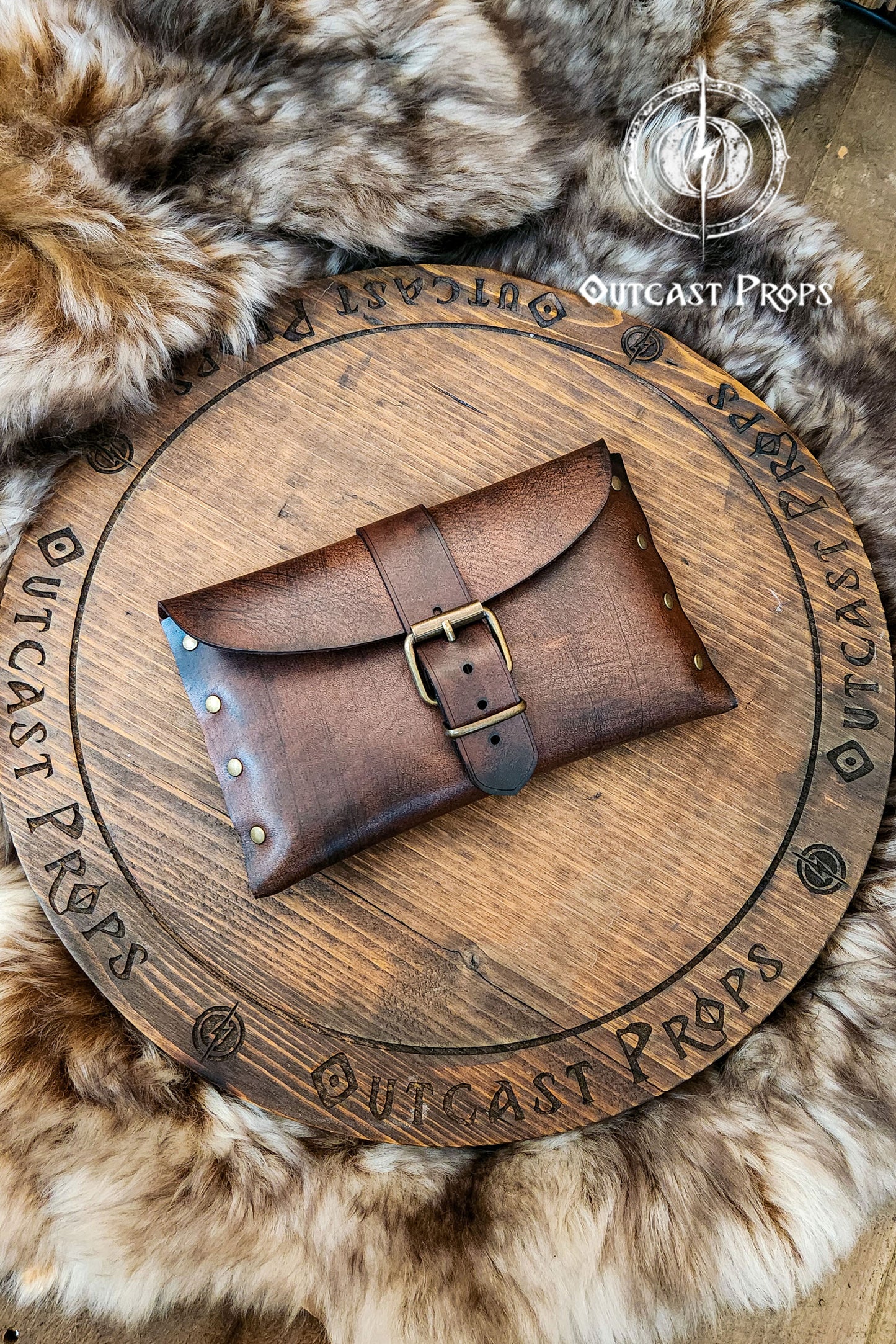 Medium brown fantasy leather belt pouch with antique brass buckle on circular wooden board engraved with Outcast Props branding. Surrounded by faux fur. Ideal for medieval scribes or LARP characters. Handcrafted pouch shows natural grain and is compatible with belts. Descriptive alt text with focus on material, use and historical style.