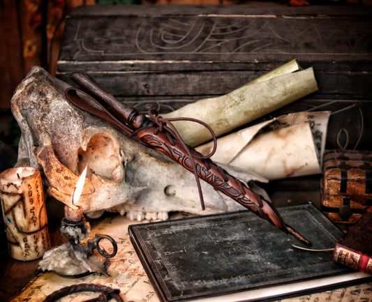 A close-up of an ornate laced leather wand holster displayed on a table with a decorative backdrop, featuring a leather-bound notebook and a candle. Magic Wand leather Holster Ornated Laced & Aged, for wizard and witch costume belt holster LARP and magic conventions and fair events