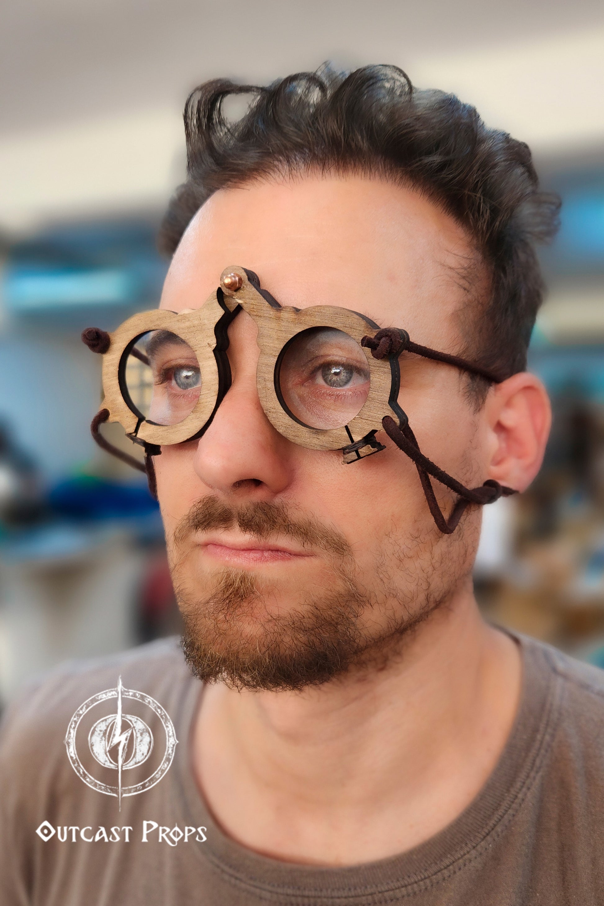 Person wearing wooden goggles with a blurred indoor background. Man wearing a wooden medieval glasses with Outcast Props logo in the corner. Medieval eyeglasses steampunk glasses larp alchemist glasses renaissance fair eyewear medieval spectacles fantasy glasses cosplay accessory. Vintage-Inspired Alchemist Spectacles: Medieval LARP Steampunk Eyewear for Renaissance Fairs & Cosplay