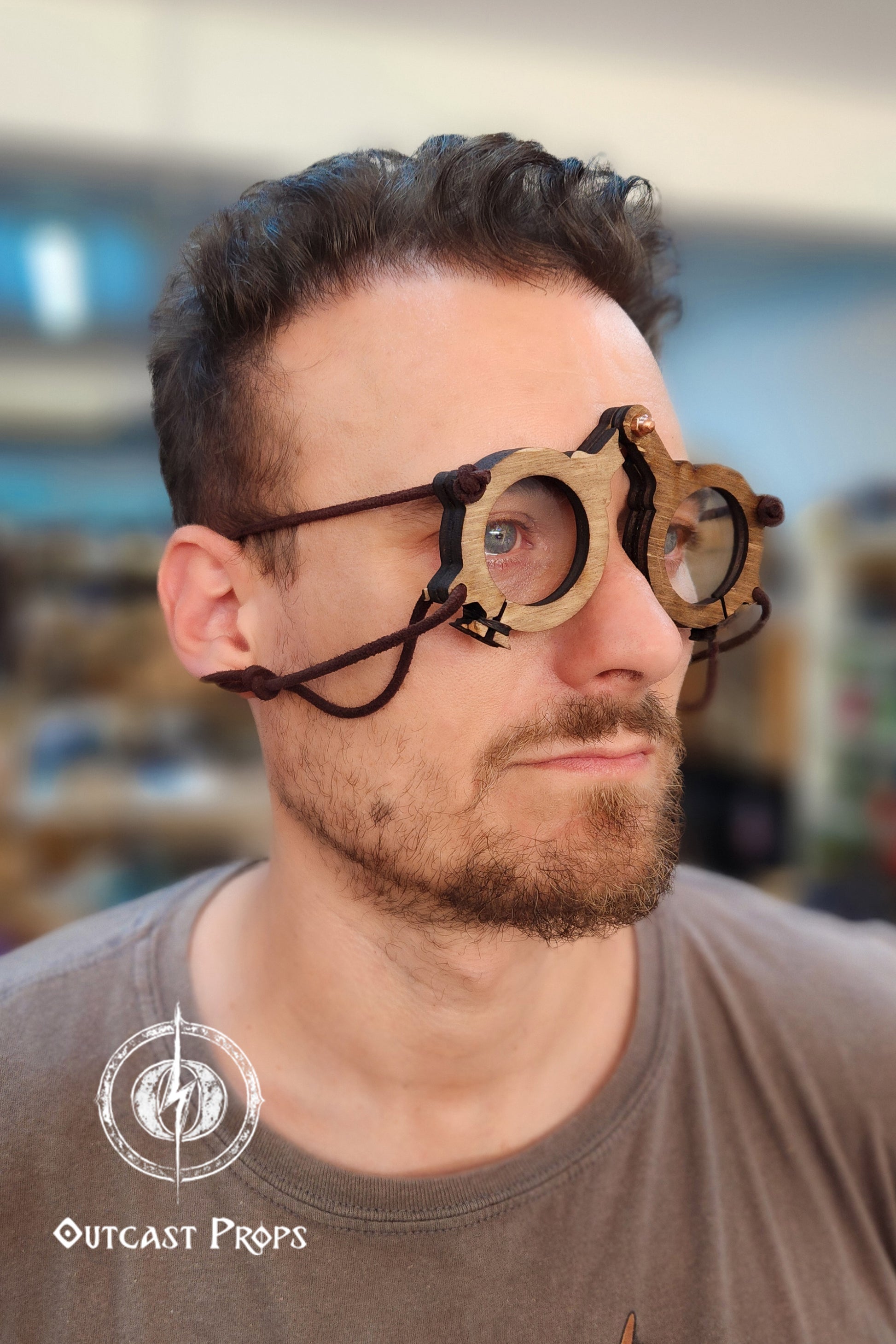 Man wearing large, round goggles with a blurred indoor background. Man wearing a wooden medieval glasses with Outcast Props logo in the corner. Medieval eyeglasses steampunk glasses larp alchemist glasses renaissance fair eyewear medieval spectacles fantasy glasses cosplay accessory. Vintage-Inspired Alchemist Spectacles: Medieval LARP Steampunk Eyewear for Renaissance Fairs & Cosplay