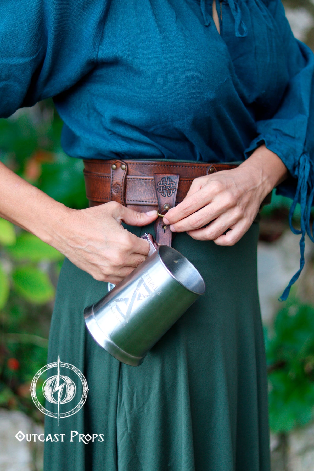Person holding a metal cup with a leather strap, wearing a green dress and blue top, against a blurred natural background. Medieval Leather Tankard Strap: Renaissance Faire Belt up to 6 cm or 2,4 inches. Accessory for key, rope, mug. Celtic or viking costume