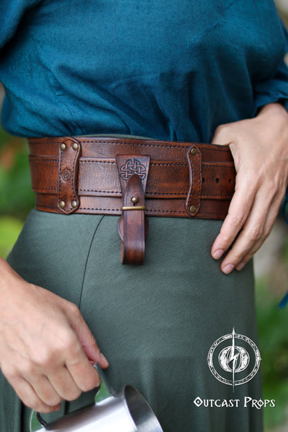 Person wearing a brown leather belt with decorative buckle, holding a sword, on a blurred background. Medieval Leather Tankard Strap: Renaissance Faire Belt up to 6 cm or 2,4 inches. Accessory for key, rope, mug. Celtic or viking costume
