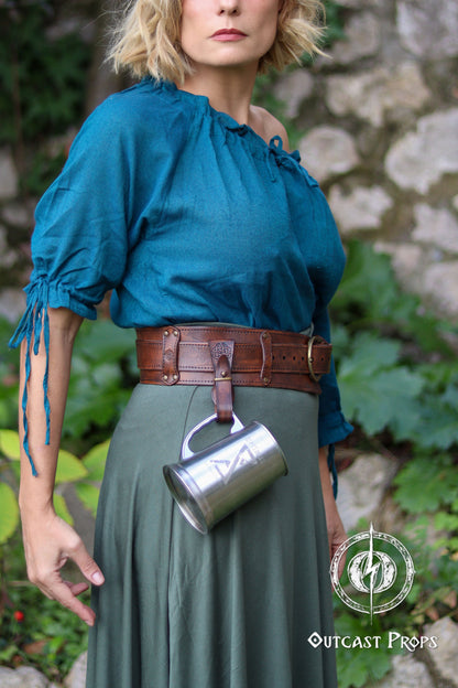 Woman wearing a blue top and green skirt with a brown belt, holding a metal cup, against a stone wall and greenery background. Medieval Leather Tankard Strap: Renaissance Faire Belt up to 6 cm or 2,4 inches. Accessory for key, rope, mug. Celtic or viking costume
