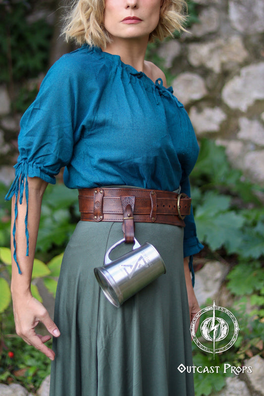 Woman wearing a blue top and green skirt with a brown belt, holding a metal cup, against a stone wall and greenery background. Medieval Leather Tankard Strap: Renaissance Faire Belt up to 6 cm or 2,4 inches. Accessory for key, rope, mug. Celtic or viking costume