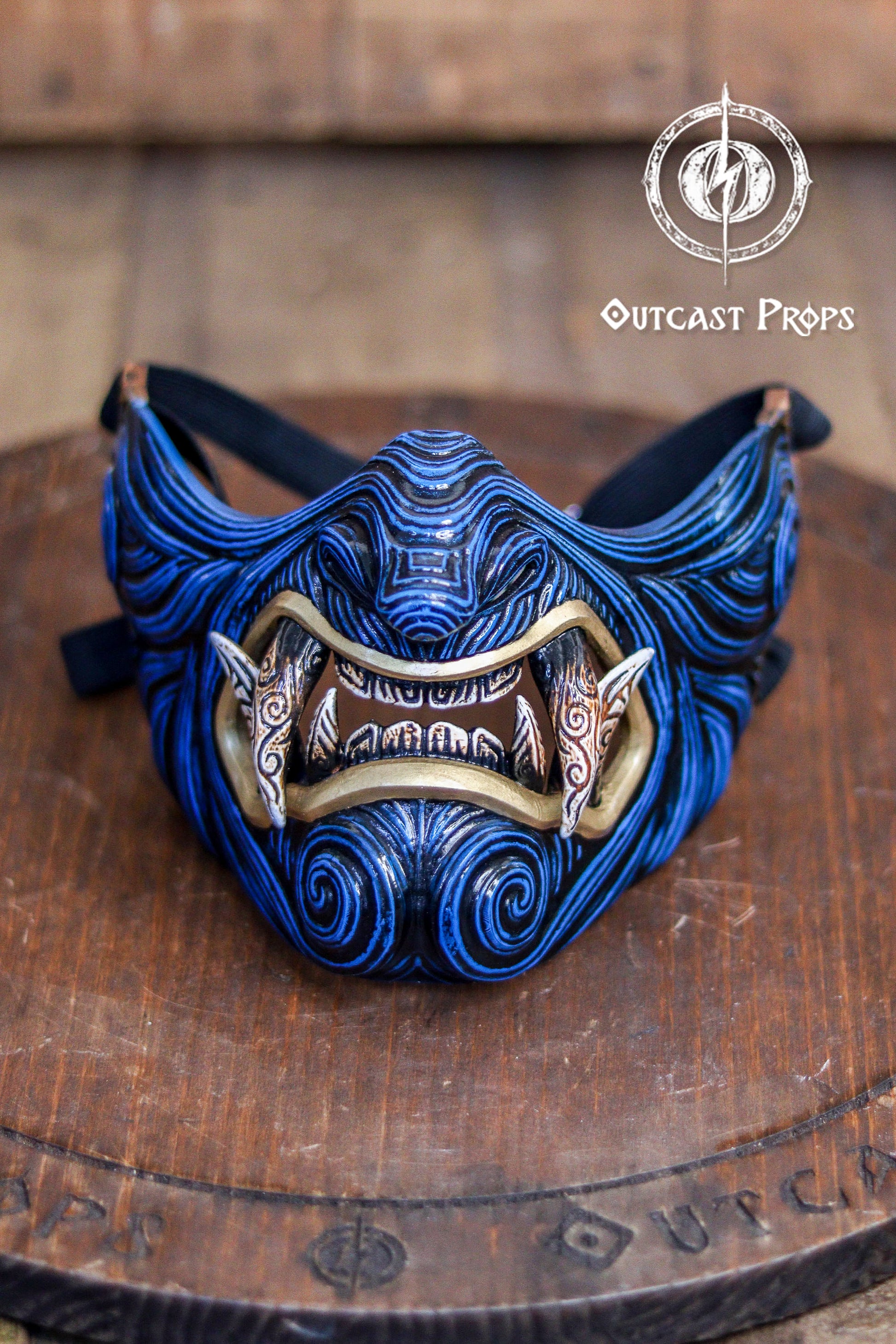 Blue and gold mask on a wooden platform with 'Outcast Props' branding. A detailed and colorful Oni Samurai Mempo mask with intricate hand-painted designs, displayed on a surface with a background of flowers and an old map. Samurai oni mask japanese half oni mask samurai wearable mask for cosplay larp menpo mask scorpion clan mask l5r samurai mask