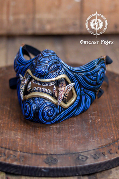 Blue and gold mask on a wooden platform with 'Outcast Props' branding. A detailed and colorful Oni Samurai Mempo mask with intricate hand-painted designs, displayed on a surface with a background of flowers and an old map. Samurai oni mask japanese half oni mask samurai wearable mask for cosplay larp menpo mask scorpion clan mask l5r samurai mask
