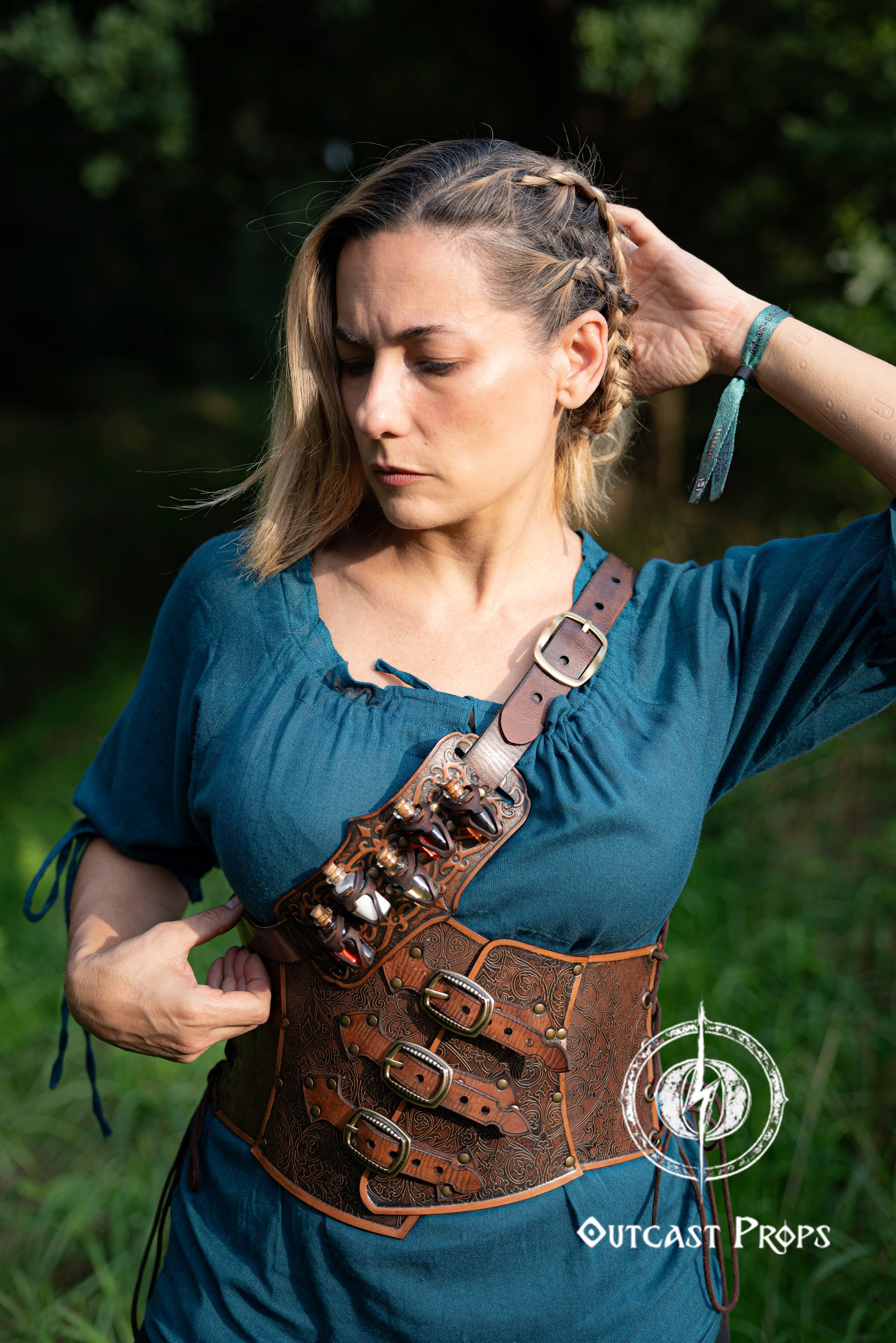 Woman wearing a leather harness in a natural setting with 'Outcast Props' branding. A handmade leather potion holder with ornate embossed patterns, designed to hold five small glass vials. The holder is displayed on a furry surface, and the vials have cork stoppers. Ornated Belt potion holder | 5  potion Belt carrier | LARP, cosplay, fantasy alchemist accessory, adventurers, steampunk, alchemist, healers