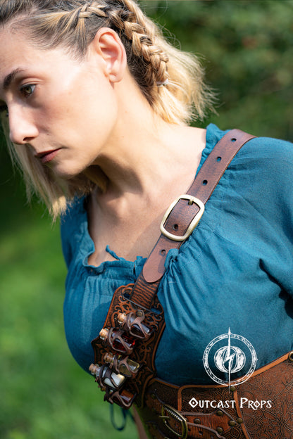Person wearing a blue top with leather straps and Outcast Props logo in the corner. A handmade leather potion holder with ornate embossed patterns, designed to hold five small glass vials. The holder is displayed on a furry surface, and the vials have cork stoppers. Ornated Belt potion holder | 5  potion Belt carrier | LARP, cosplay, fantasy alchemist accessory, adventurers, steampunk, alchemist, healers