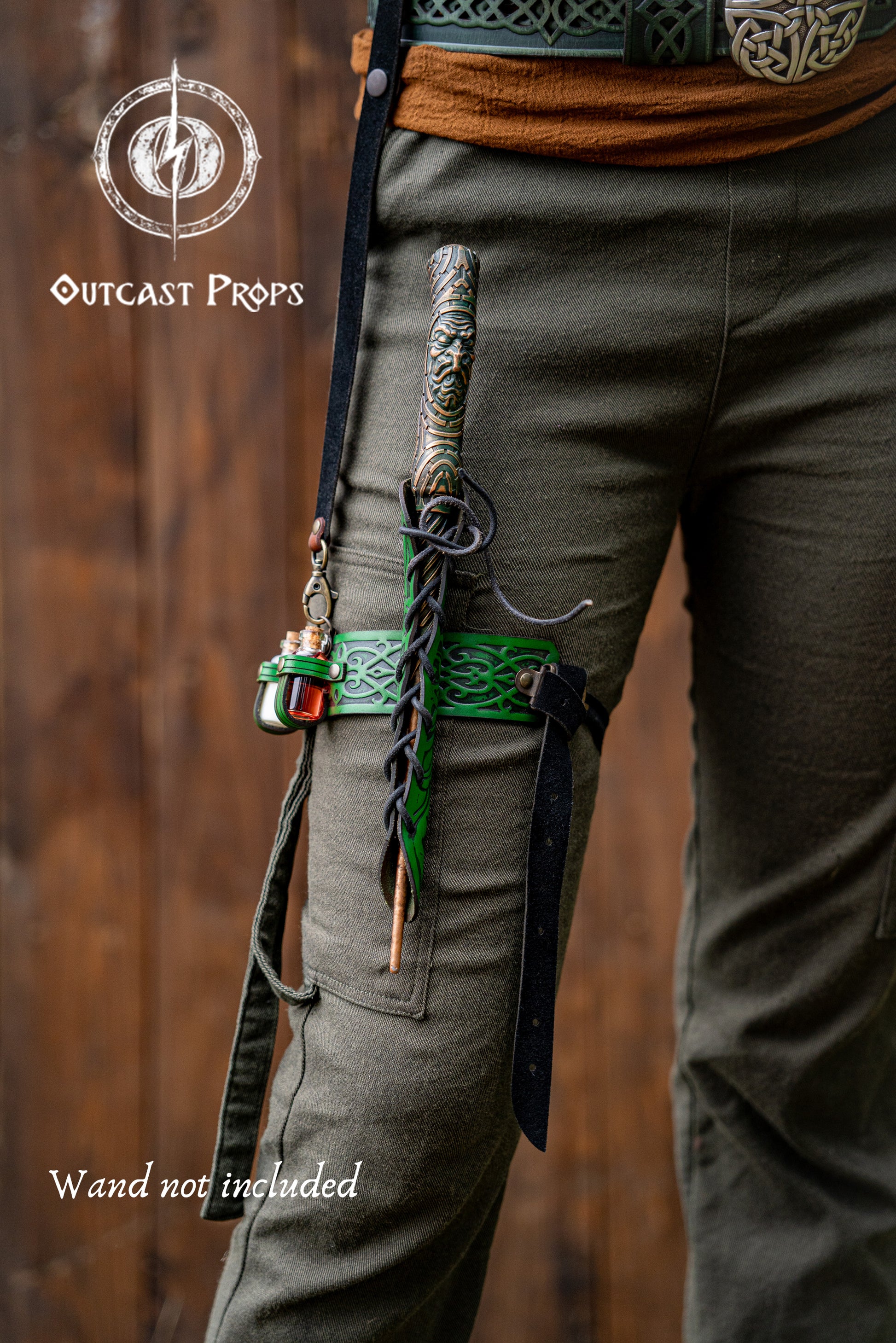 Person wearing green pants with a sword on a wooden background, featuring 'Outcast Props' branding. Cosplayer wears a green embossed leather wand holster garter with potion bottles and a rustic fantasy look. The wand holster wraps around the thigh, secured by adjustable straps. A perfect cosplay wand holder for witches, druids, or mages. The holster doubles as a larp potion holster, keeping tools of magic close. Designed for immersive fantasy costumes and magic users who attend LARP, Ren Faires or wizard