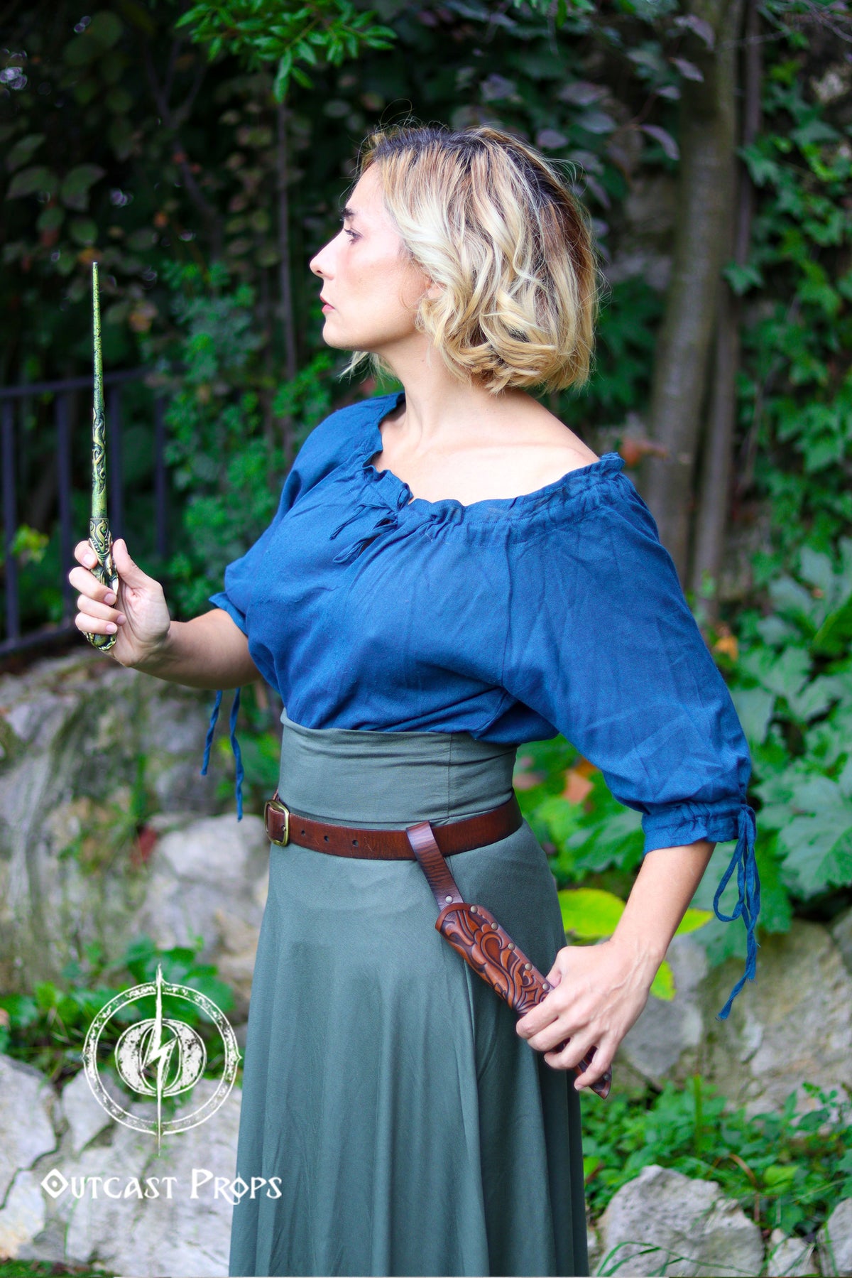 A blonde woman dressed in a teal blouse and long green skirt stands in a garden holding a decorative wand, while at her hip hangs a brown embossed leather wand holster secured to a belt. The holster features ornate carved patterns and an open tip that lets the wand protrude. Surrounded by greenery and stone, the scene evokes a wizard or elf mage at a LARP or Renaissance faire. The vegetable tanned leather sheath adds a handcrafted fantasy touch suitable for witch cosplay or dark wizard characters.