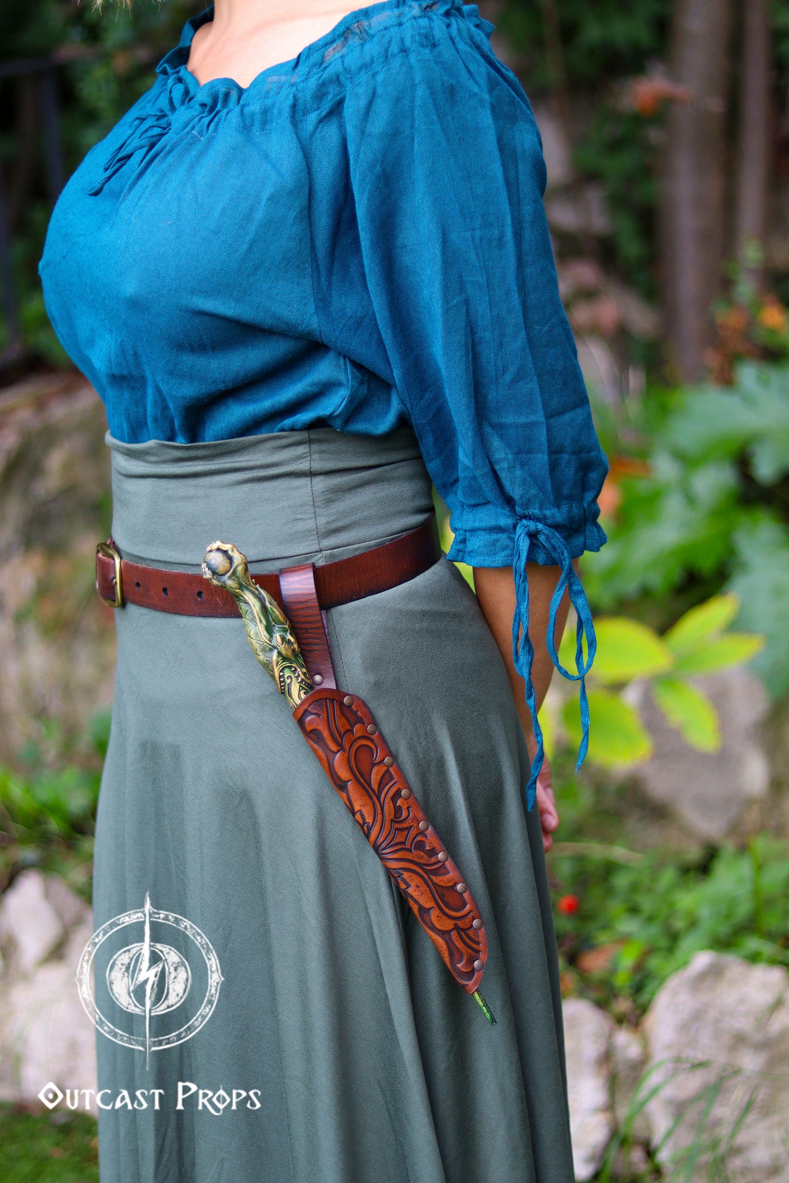 Side view of a woman in a teal blouse and flowing green skirt wearing a brown embossed leather wand sheath on her belt. A sculpted fantasy wand rests securely inside the holster, which shows detailed tooling and riveted construction. The belt loop keeps the holder close to the hip for easy access, blending seamlessly into a wizard, witch, elf mage or dark wizard costume. The outdoor setting with foliage creates an immersive LARP or ren faire atmosphere, highlighting the handcrafted leatherwork.