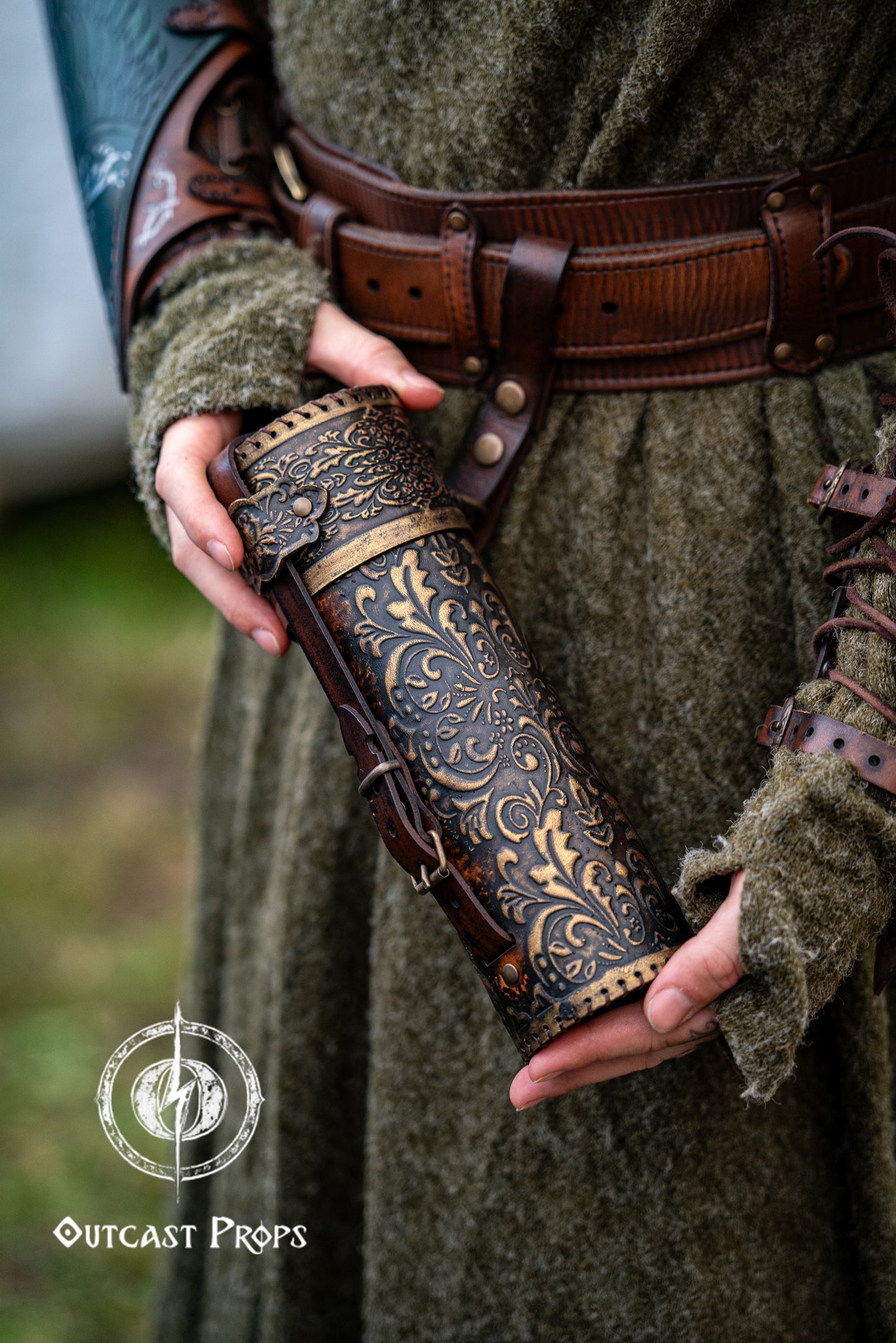 Person holding a decorative leather weapon sheath with intricate patterns, wearing a green outfit. A brown leather scroll case with golden embossed floral patterns is held by a person wearing a textured green medieval tunic and wide fantasy belt. Perfect accessory for LARP wizards, fantasy scholars, or noble messengers. Handmade with vegetable-tanned leather, ideal for cosplay, historical reenactment, and Renaissance fair characters. Elegant leather tube for scrolls or secret maps.