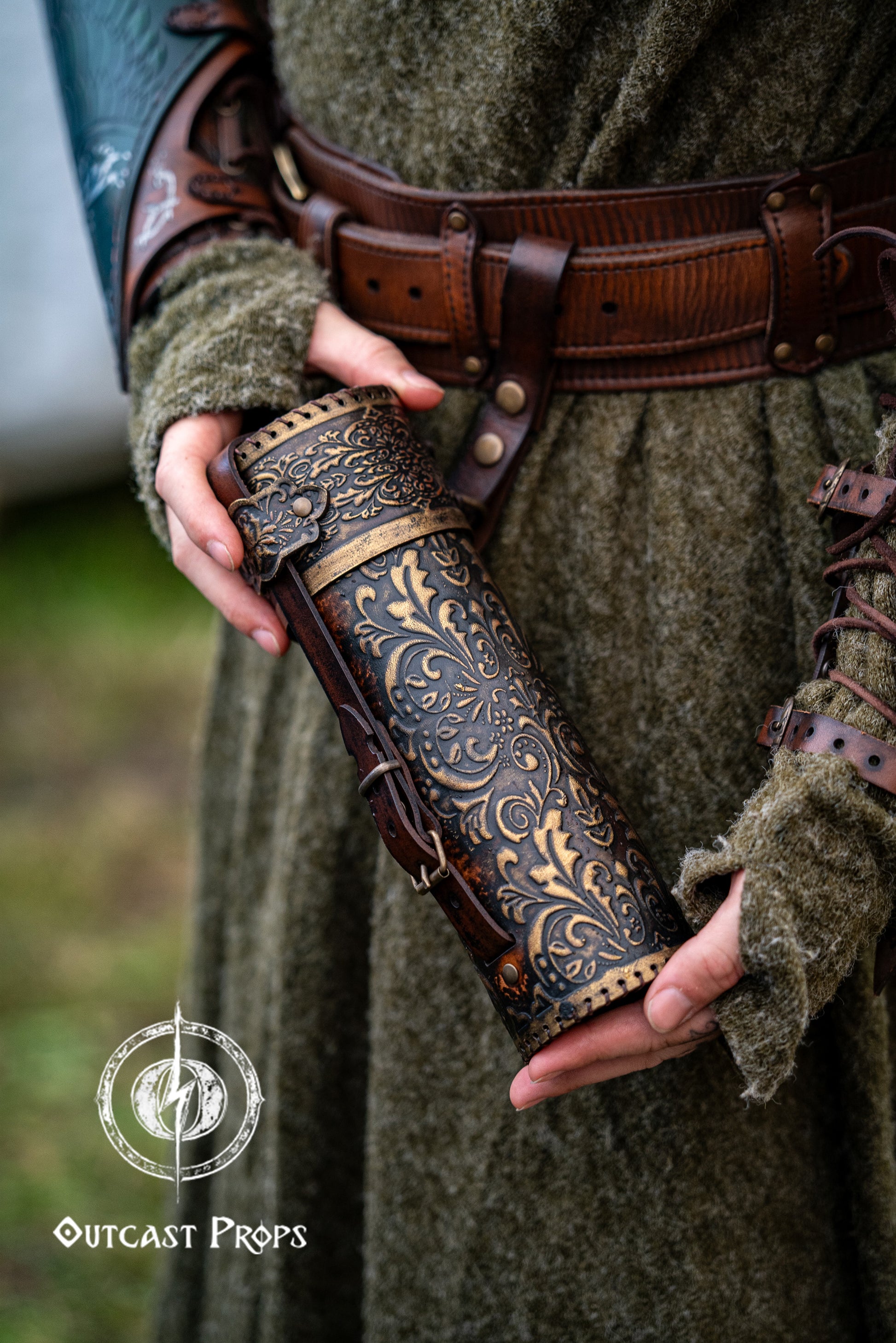 Person holding a decorative leather weapon sheath with intricate patterns, wearing a green outfit. A brown leather scroll case with golden embossed floral patterns is held by a person wearing a textured green medieval tunic and wide fantasy belt. Perfect accessory for LARP wizards, fantasy scholars, or noble messengers. Handmade with vegetable-tanned leather, ideal for cosplay, historical reenactment, and Renaissance fair characters. Elegant leather tube for scrolls or secret maps.