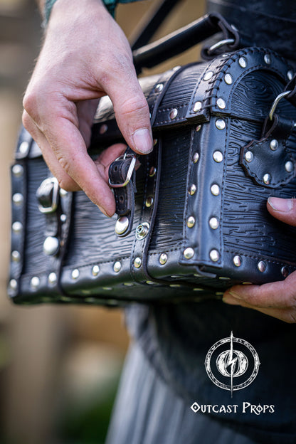 Person holding a black leather bag with studs outdoors, featuring 'Outcast Props' logo. Person wearing a dark outfit with a leather bag, holding a belt with multiple buckles. Man wearing a pirate chest leather bag with studs, standing outdoors. Leather treasure chest pirate chest bag larp pirate backpack treasure chest purse pirate accessories cosplay renfaire pouch belt bag