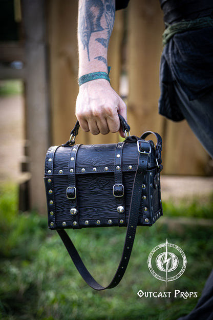 Person holding a black leather bag with studs outdoors, featuring 'Outcast Props' logo. Person wearing a dark outfit with a leather bag, holding a belt with multiple buckles. Man wearing a pirate chest leather bag with studs, standing outdoors. Leather treasure chest pirate chest bag larp pirate backpack treasure chest purse pirate accessories cosplay renfaire pouch belt bag