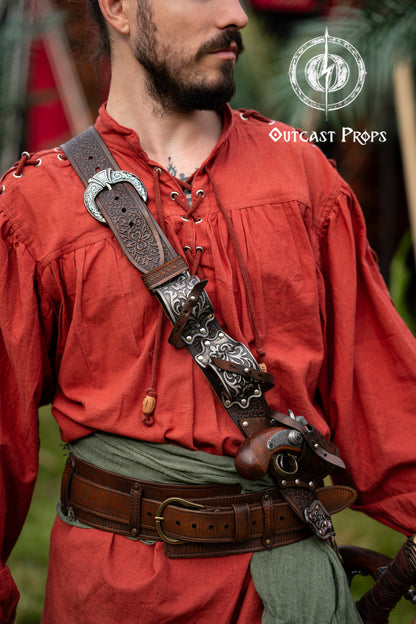 Close-up of pirate wearing silver-engraved leather baldric inspired by Blackbeard and Anne Bonny. Holds flintlock pistols and sword, perfect for LARP, pirate cosplay or Renaissance fairs. Premium handmade belt crafted with vegetable-tanned leather. Ideal for Jack Sparrow costumes, Pirate Weekend and themed events. High detail on buckles, engravings and leather texture. Person wearing a red outfit with a decorative leather strap and Outcast Props logo.