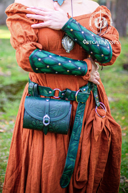 A person wearing a medieval-style costume dress in rust orange, with green leather arm guards, a matching green belt and pouch, and a silver pendant. The photo is taken outdoors on grass, with a visible 'Outcast Props' watermark.