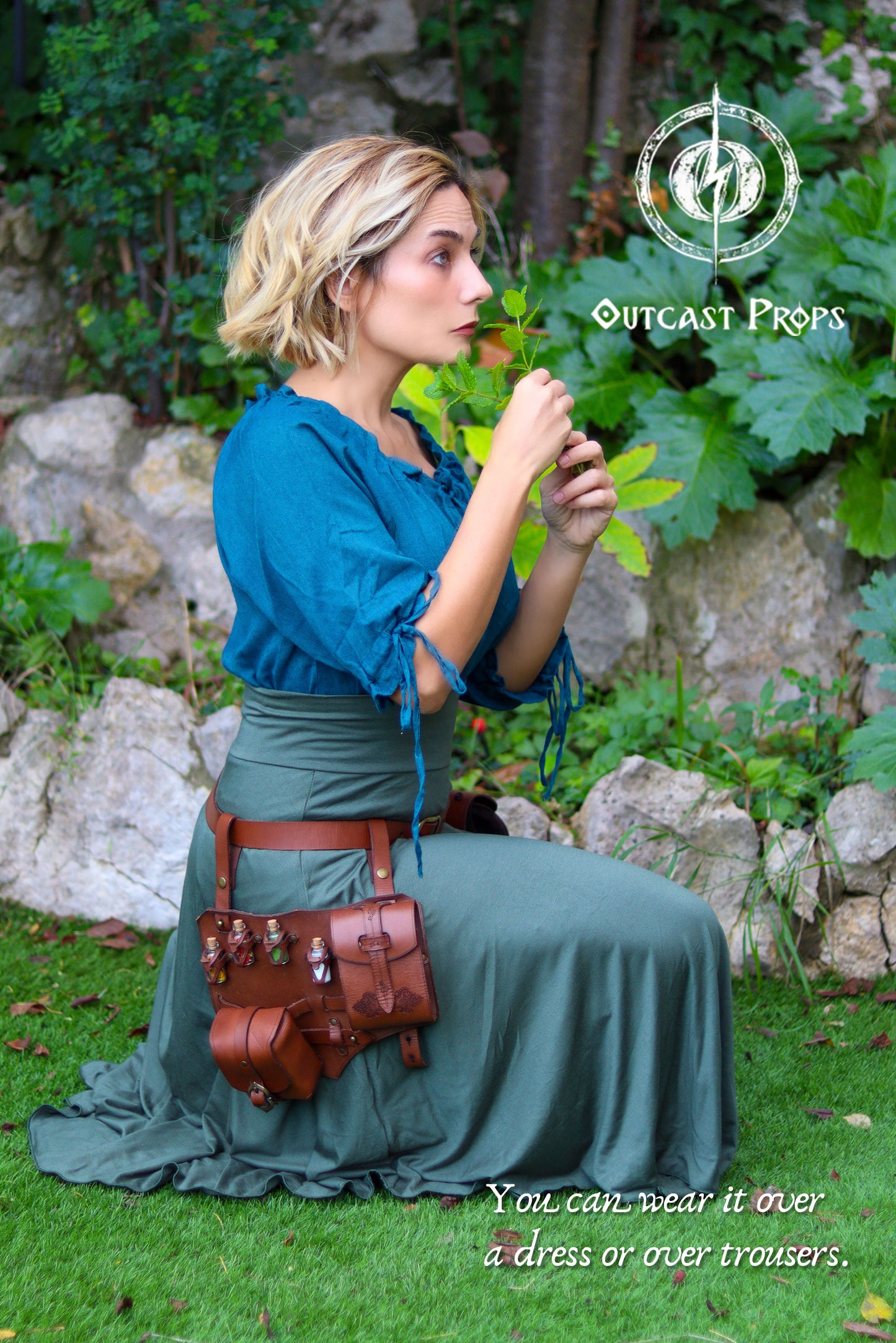 A blonde woman kneels in a garden wearing a teal blouse and long green skirt while smelling a fresh leaf, styled like an herbalist or alchemist at a LARP or Ren Faire. At her hip hangs a brown leather potion belt with small vials, a pouch and a book holster style pocket, made with aged artisan leather. The leg harness sits smoothly over the skirt, showing how the potion belt can be worn over dresses or trousers for steampunk or larp alchemist costumes.