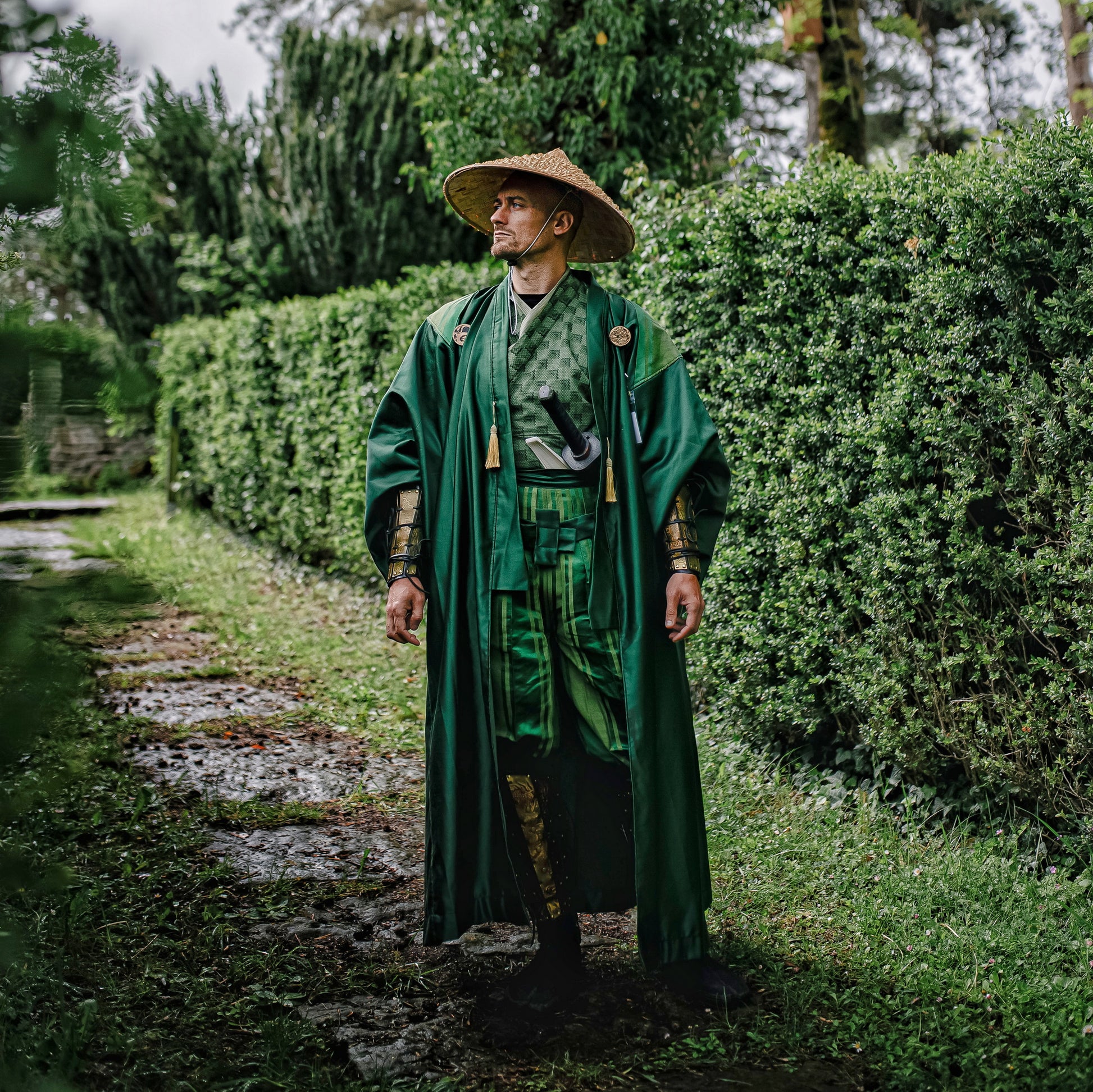 Person dressed in a green Japanese-inspired samurai outfit, wearing leather shin armor with golden dragon engravings, standing on a garden path surrounded by lush greenery. Costume includes matching bracers and robe, perfect for samurai cosplay and LARP characters.