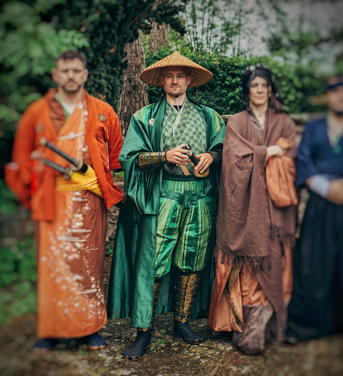 Three people dressed in Japanese-themed fantasy costumes, with the central figure wearing a green robe, striped hakama trousers, and golden leather shin armor with dragon embossing. A striking example of full samurai cosplay for LARP or themed events.