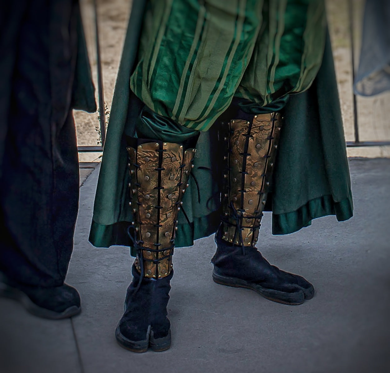Close-up of the lower legs of a cosplayer in green robes, showing leather samurai greaves with detailed gold dragon patterns. Worn over black tabi-style shoes, the shin armor is laced securely and forms part of a complete fantasy samurai outfit.