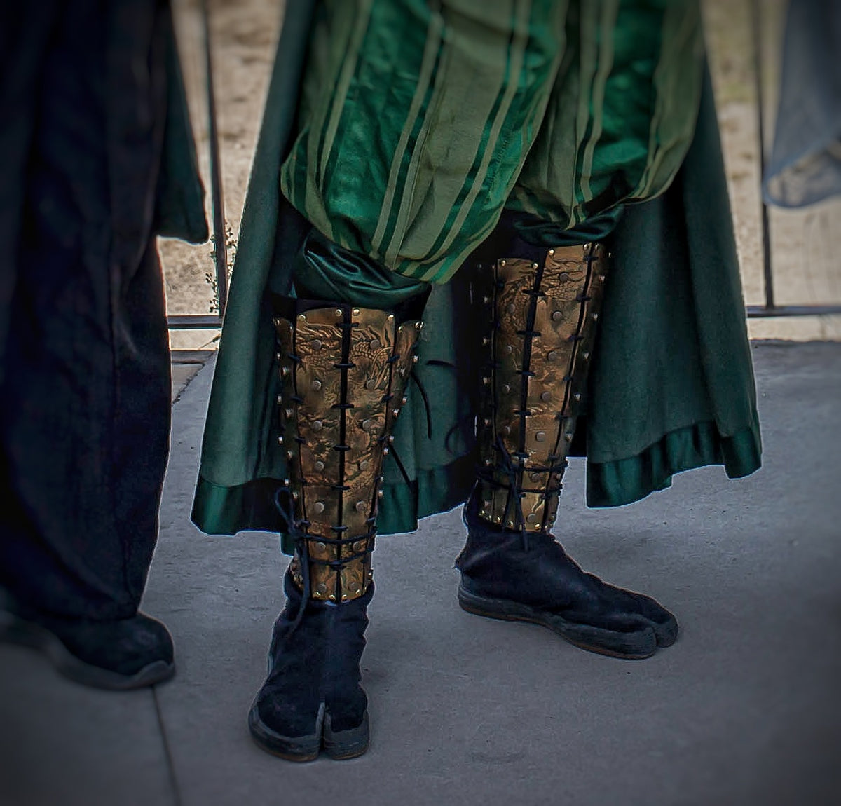 Close-up of the lower legs of a cosplayer in green robes, showing leather samurai greaves with detailed gold dragon patterns. Worn over black tabi-style shoes, the shin armor is laced securely and forms part of a complete fantasy samurai outfit.