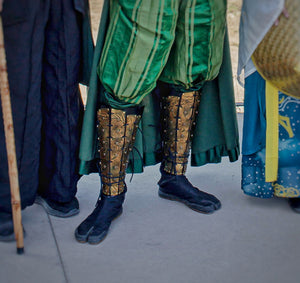 Zoom on greaves with embossed oriental dragon patterns worn over black split-toe boots, part of a green samurai costume for fantasy events or LARP. The leather armor is hand-dyed and riveted, designed for durability and visual impact.