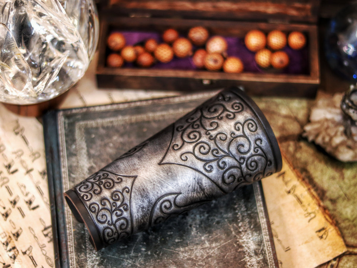 A pair of silver leather bracers with a decorative pattern, displayed on a surface with a backdrop of a mirror, books, and other antiquities. Shadowbinder bracer for mages & wizards, sorcerer vambarce for larp costume and wizardry cosplay leather witchcraft handmade